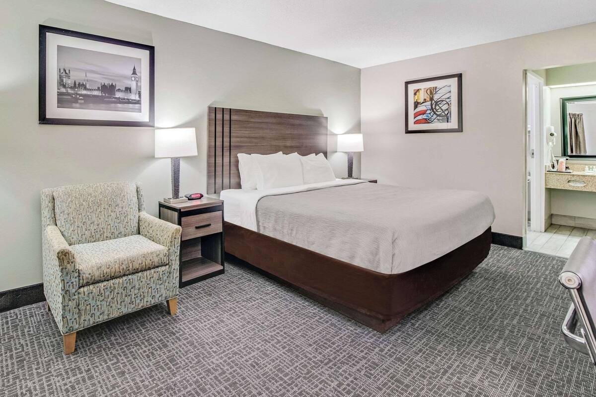 Una habitación de hotel con una cama grande, una silla, una mesita de noche y dos lámparas de pared.