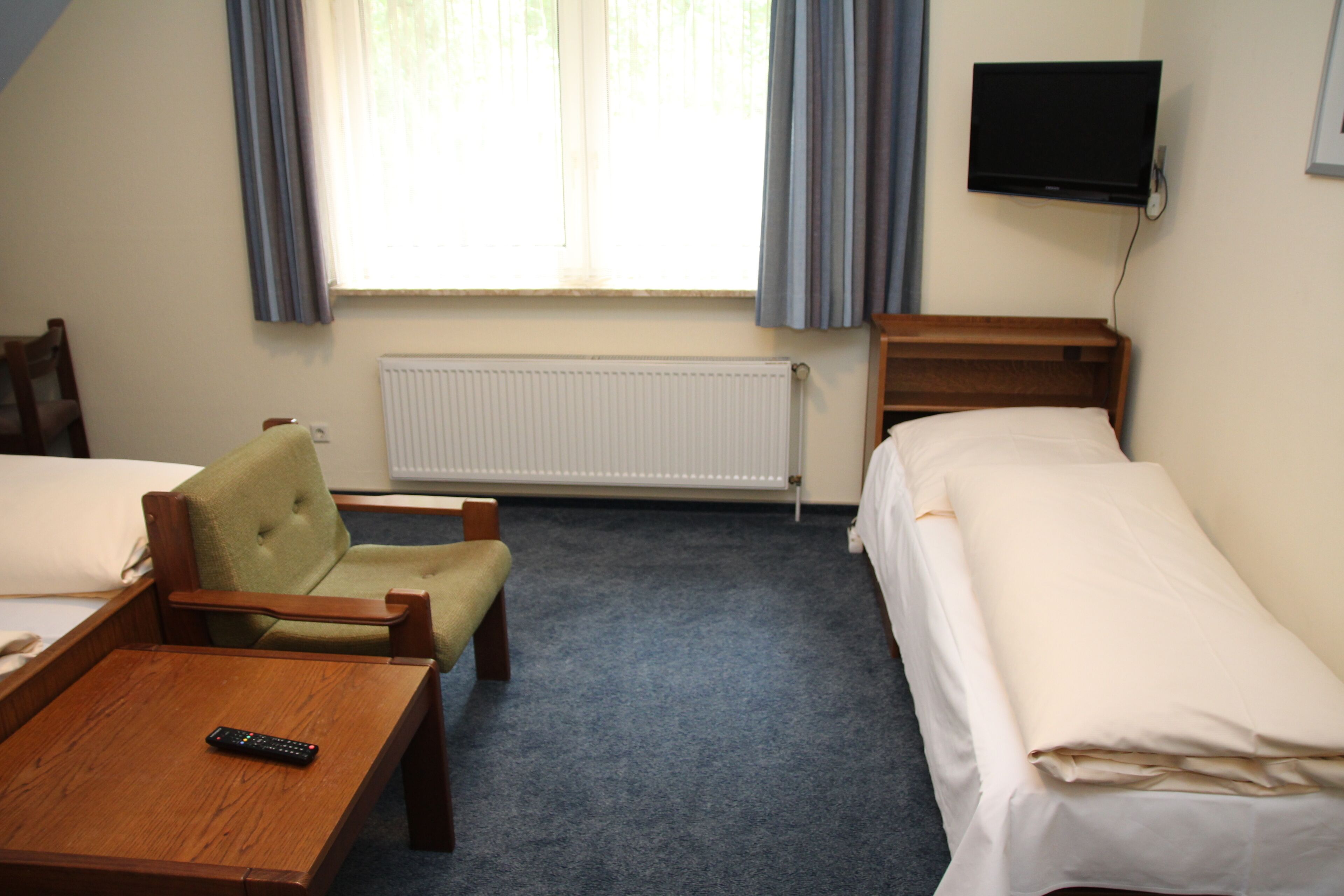 Habitación de hotel con una cama individual, una silla, una mesita, televisión y ventana con cortinas.
