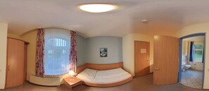 Un dormitorio con cama, mesita de noche con lámpara, radiador, ventana con cortinas, puerta y baño a la vista a través de una puerta abierta.