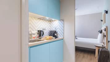 A compact kitchen with a sink, coffee maker, and a bag of snacks. A bed with a pillow and a suitcase is visible in the adjacent room.