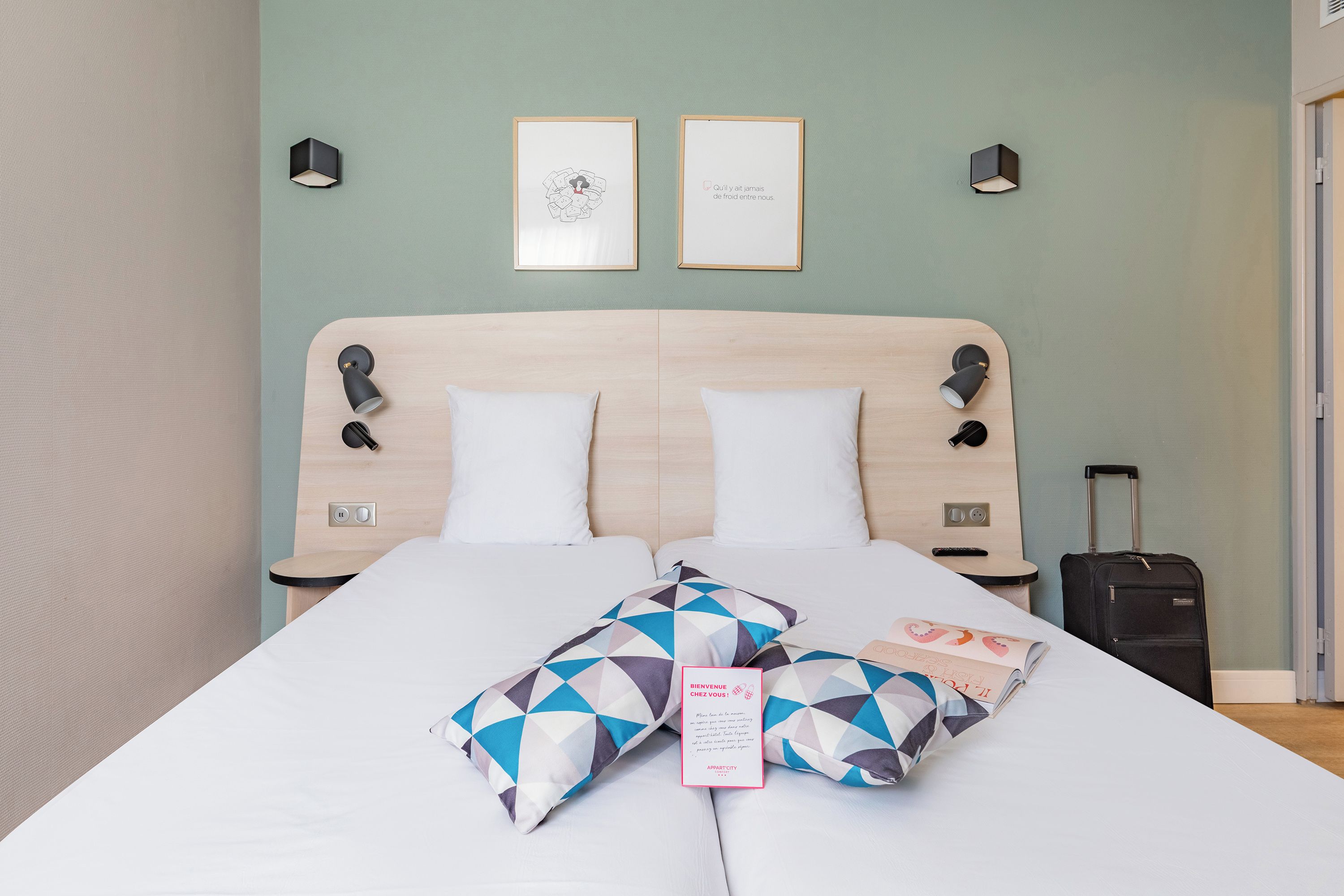 A hotel room with a bed, two wall-mounted lamps, a framed artwork, and a suitcase.