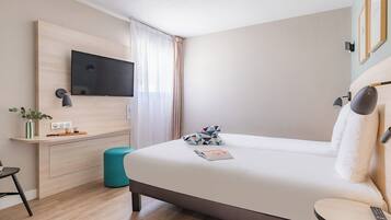 A modern hotel room with a large bed, a TV mounted on the wall, a bedside table with a remote, and a small desk area with a lamp and a plant.