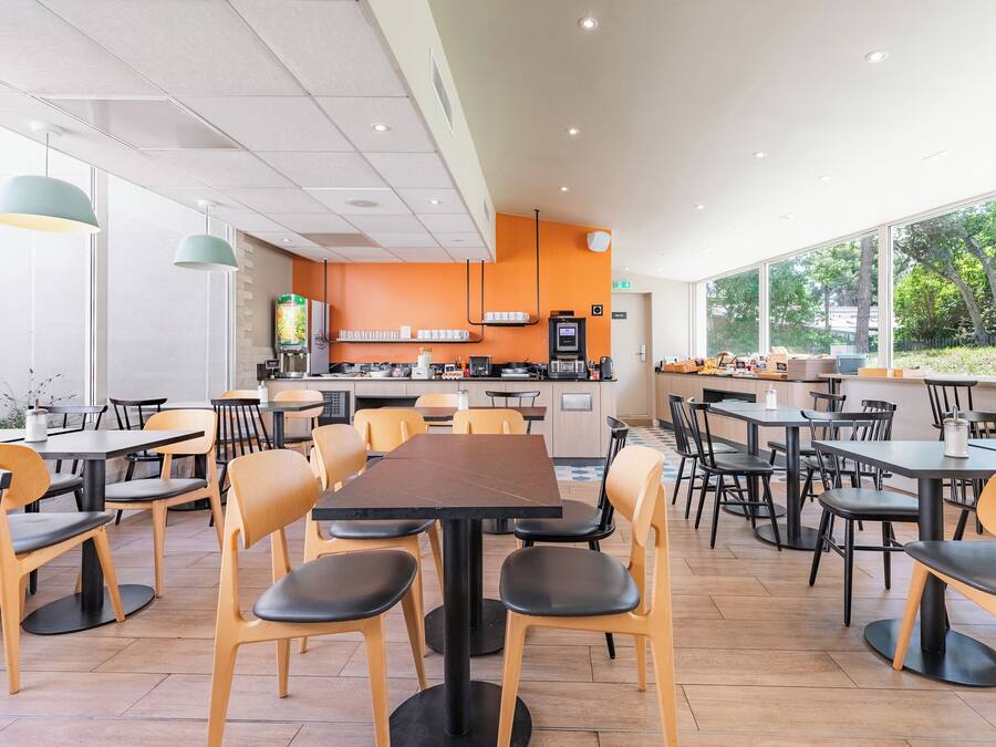 A modern cafe with multiple tables and chairs, a kitchen area, and large windows offering a view of the outdoors.