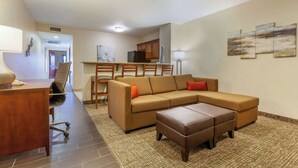 A modern living room with a sofa, ottoman, and a desk with a chair. There is a kitchen area in the background with cabinets and a refrigerator. - Comfort Suites Glendale - State Farm Stadium Area (Glendale)