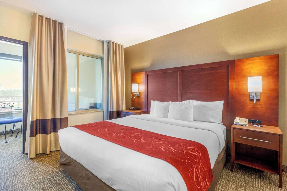 A hotel room with a large bed, a nightstand with a phone and lamp, a window with curtains, and a view of the outdoors.