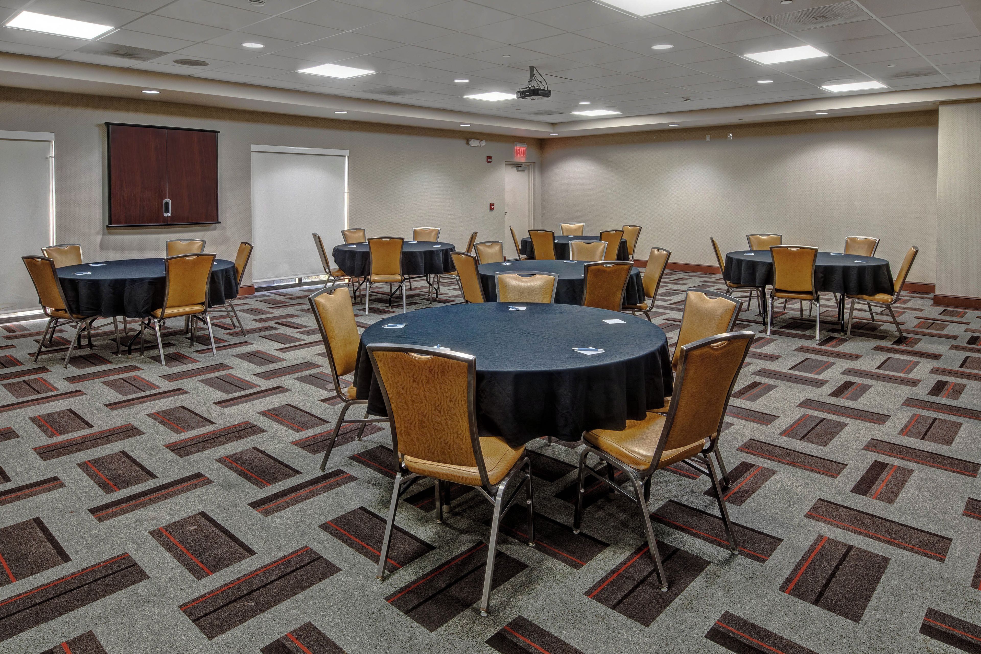 A conference room with multiple tables and chairs, carpeted floor, and a projector on the ceiling.