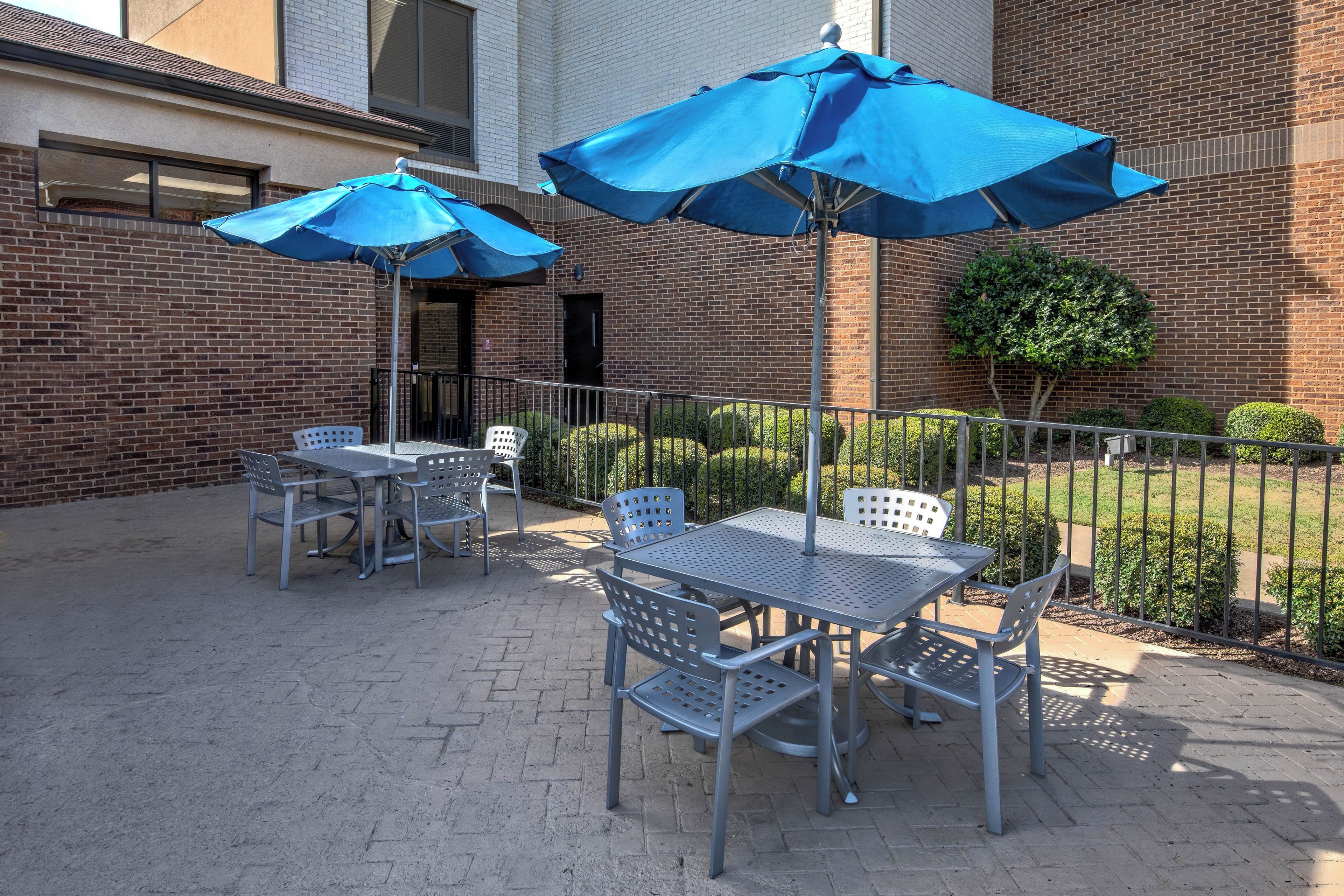 An outdoor patio with metal tables and chairs, blue umbrellas, and a brick building in the background.