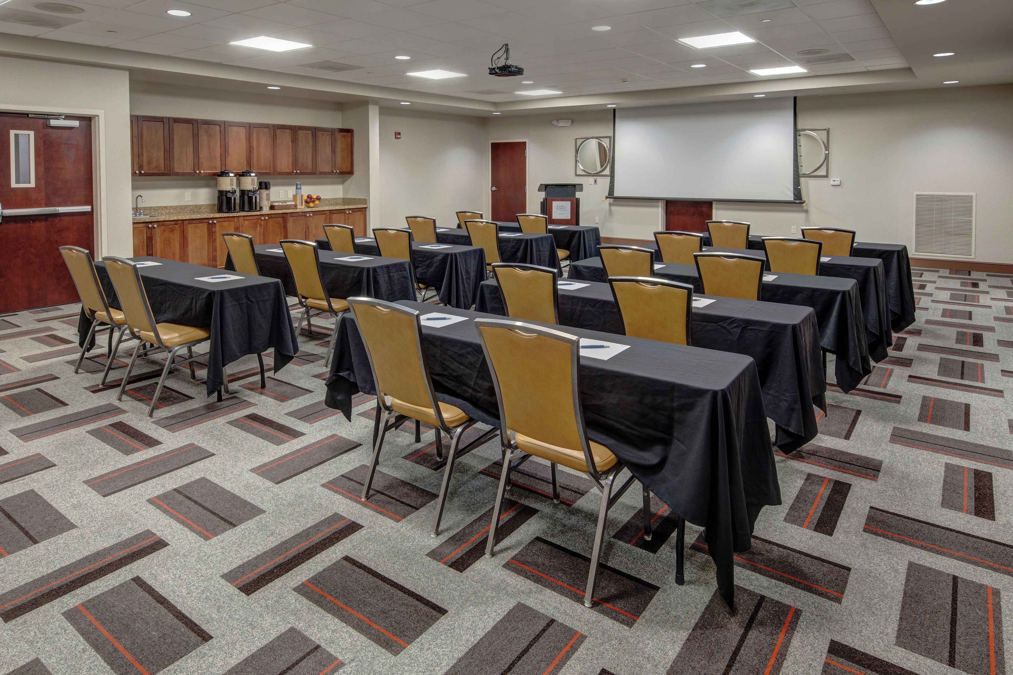 Une salle de conférence équipée de tables et de chaises, d’un écran de projection et d’un coin cuisine.