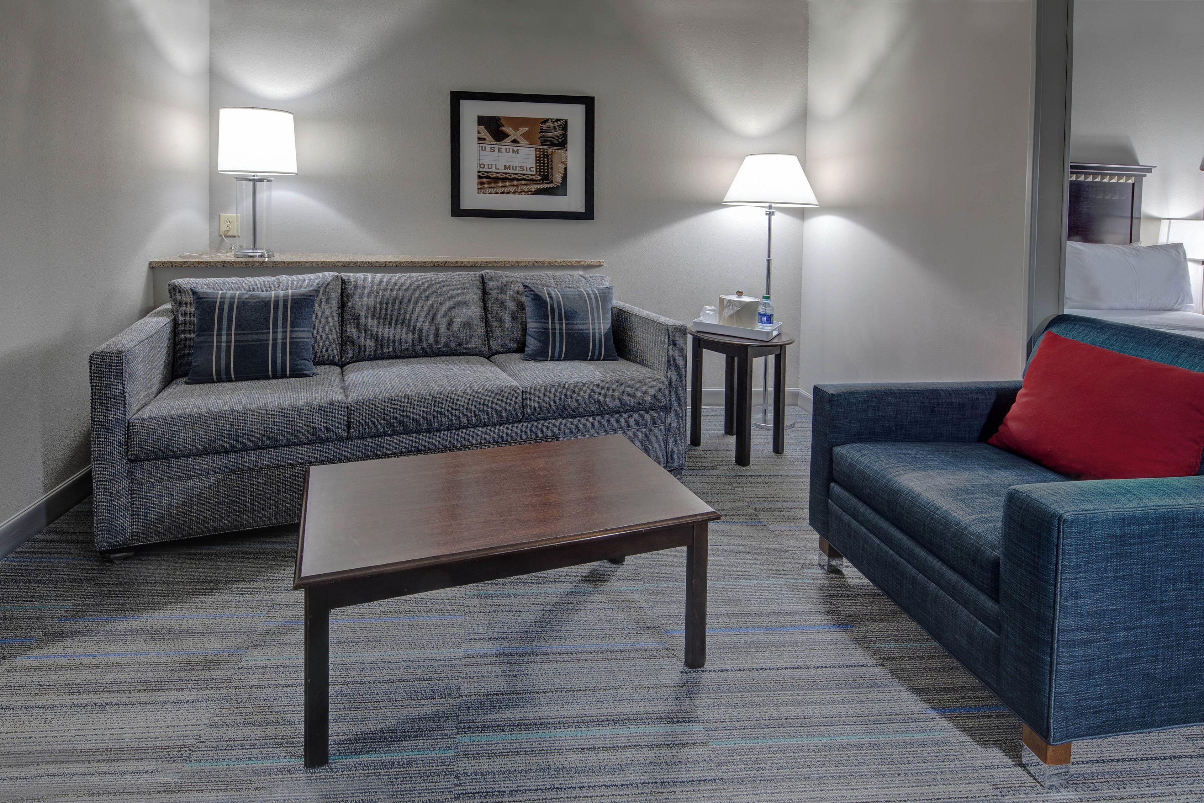 A living room with a sofa, a coffee table, and a chair. There is a framed picture on the wall and a lamp on a side table.