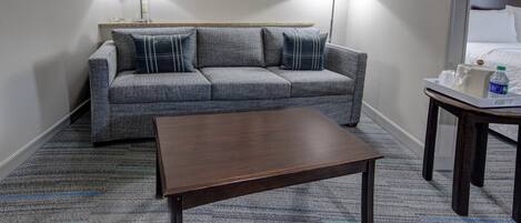 A hotel room with a grey patterned sofa, a wooden coffee table, and a small side table with a bottle.