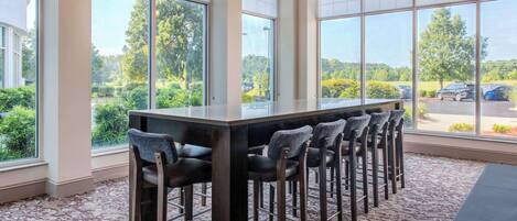 A bar area with a long table and multiple bar stools, large windows with a view of a parking lot and greenery outside.