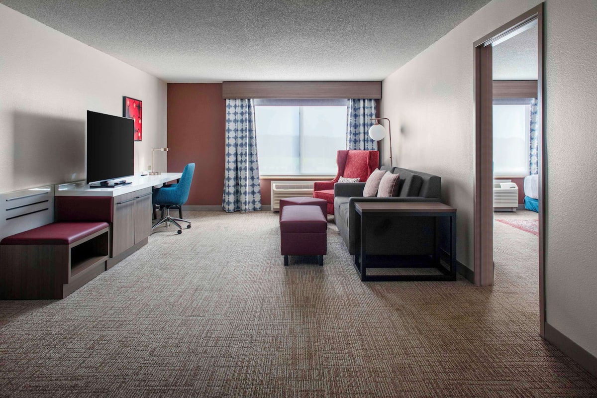 A hotel room with a desk, chair, TV, sofa, ottoman, and a window with curtains.