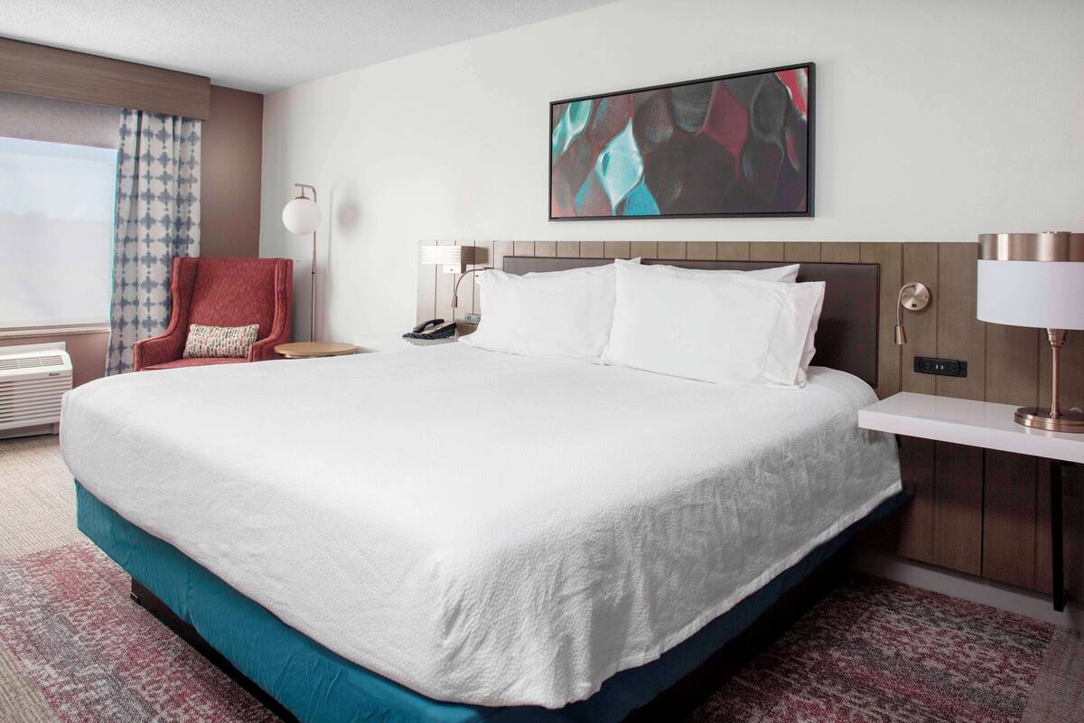 A hotel room with a large bed, a chair, a desk, and a window with curtains.