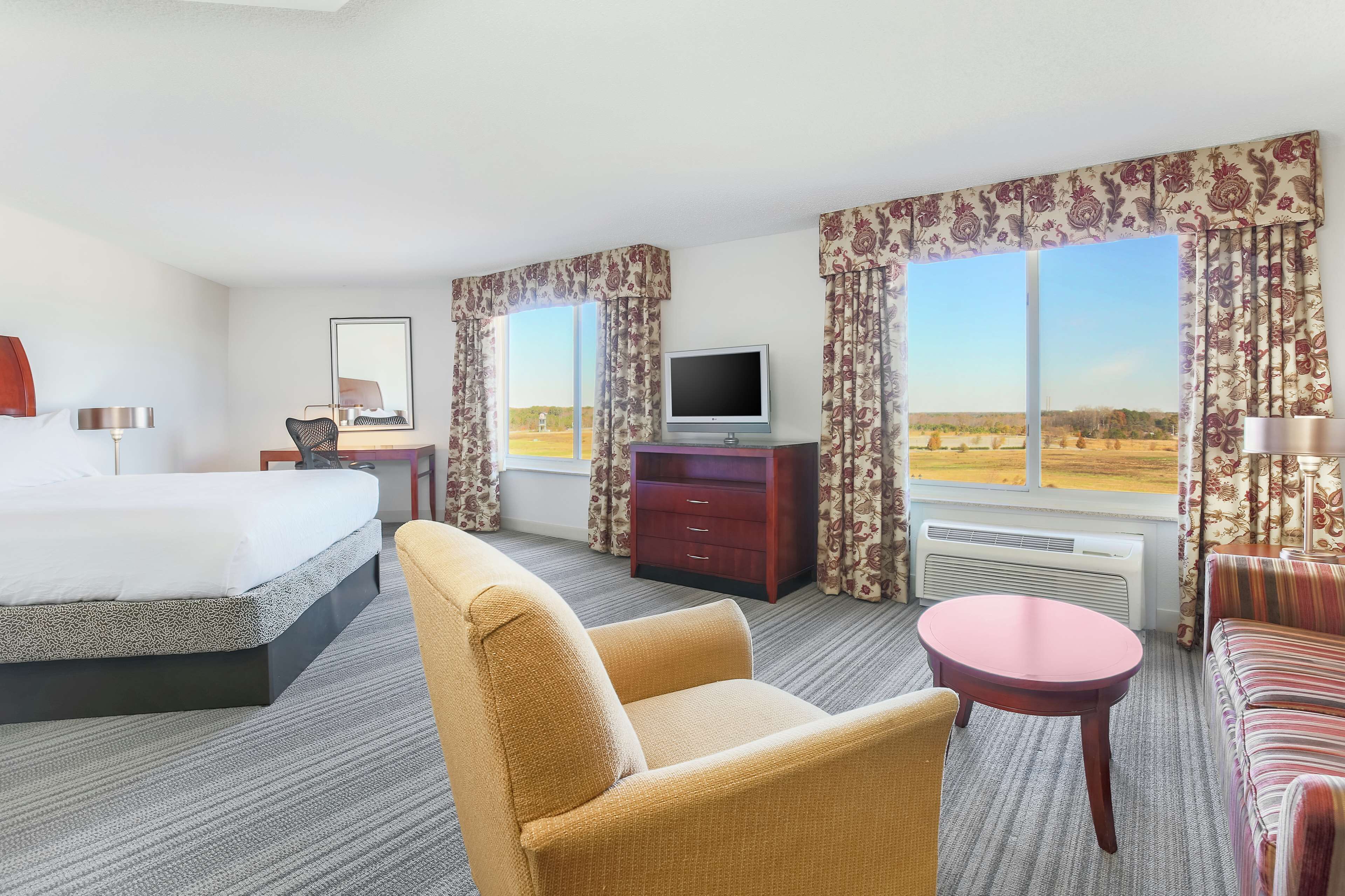 Une chambre d’hôtel comprenant un lit, un bureau, une télévision, une chaise, un canapé et de grandes fenêtres avec des rideaux.