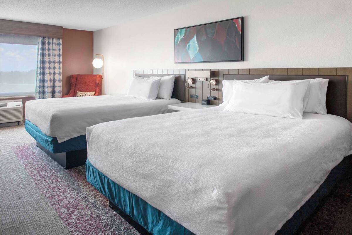 A hotel room with two beds, a large window with curtains, a flat-screen TV, and a wall-mounted light fixture.