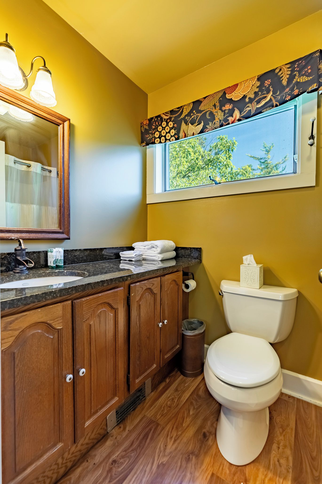 A bathroom with wooden cabinets, a white toilet, a window with a view of trees, and a mirror with lights.
