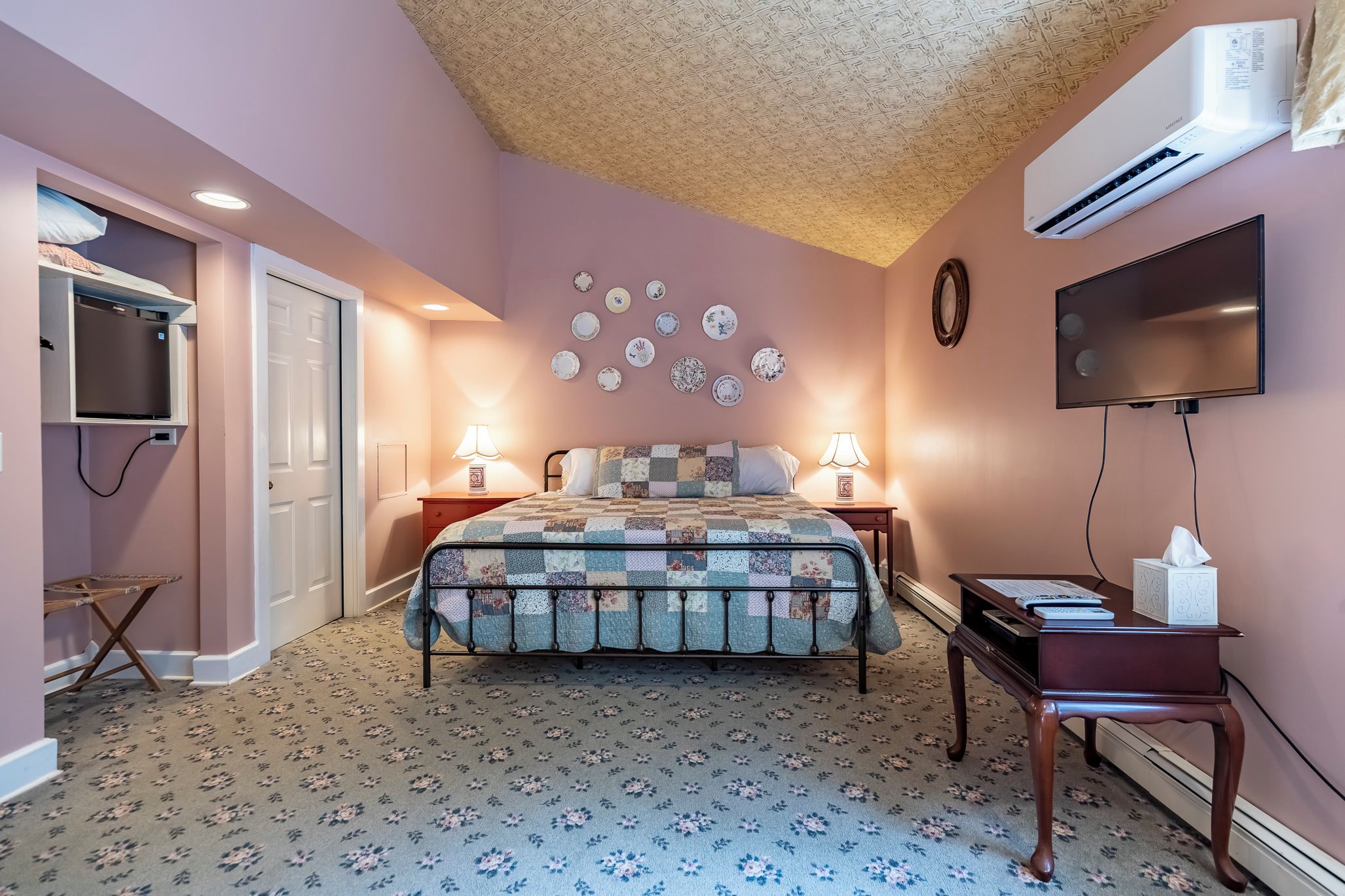 A bedroom with a bed, bedside lamps, a flat-screen TV, a wall-mounted air conditioner, and a decorative plate collection.