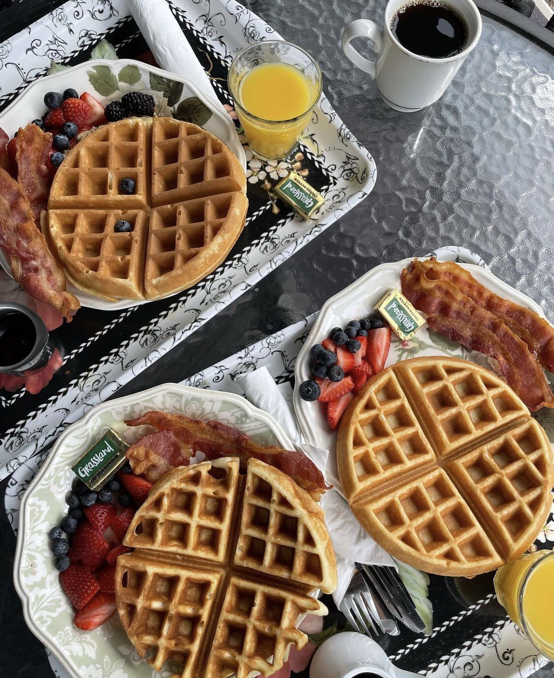 Dos platos con gofres, fresas, arándanos y tocino, junto con un vaso de jugo de naranja y una taza de café.