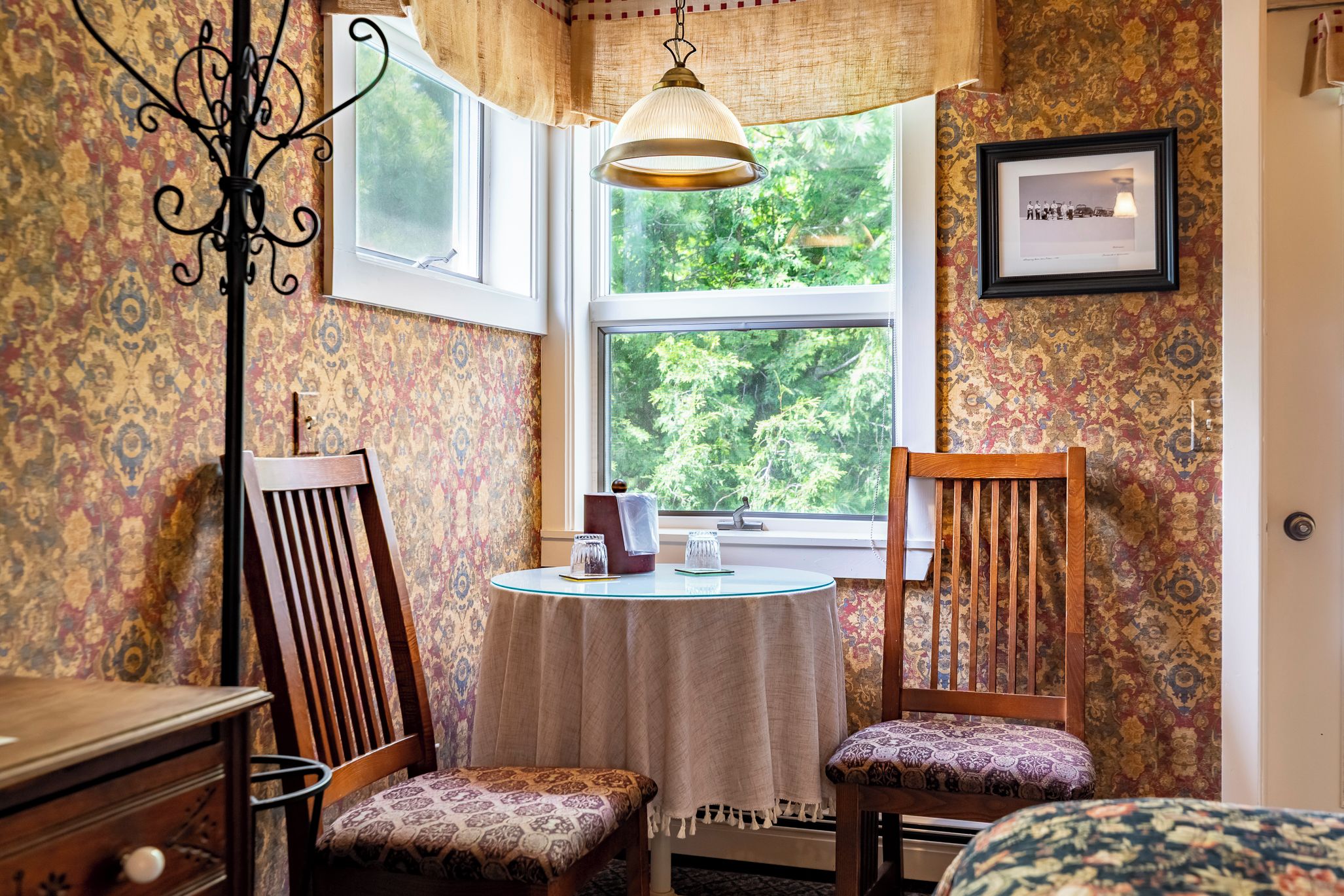 A room with a round table, two chairs, a hanging light fixture, a window with a view of greenery, and patterned wallpaper.