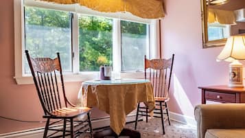 A dining area with a table and two chairs, a window with a view of trees, and a lamp on a nightstand.