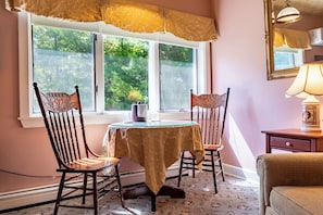 A dining area with a table and two chairs, a window with a view of trees, and a lamp on a nightstand. - The Sylvan Inn (Glen Arbor)