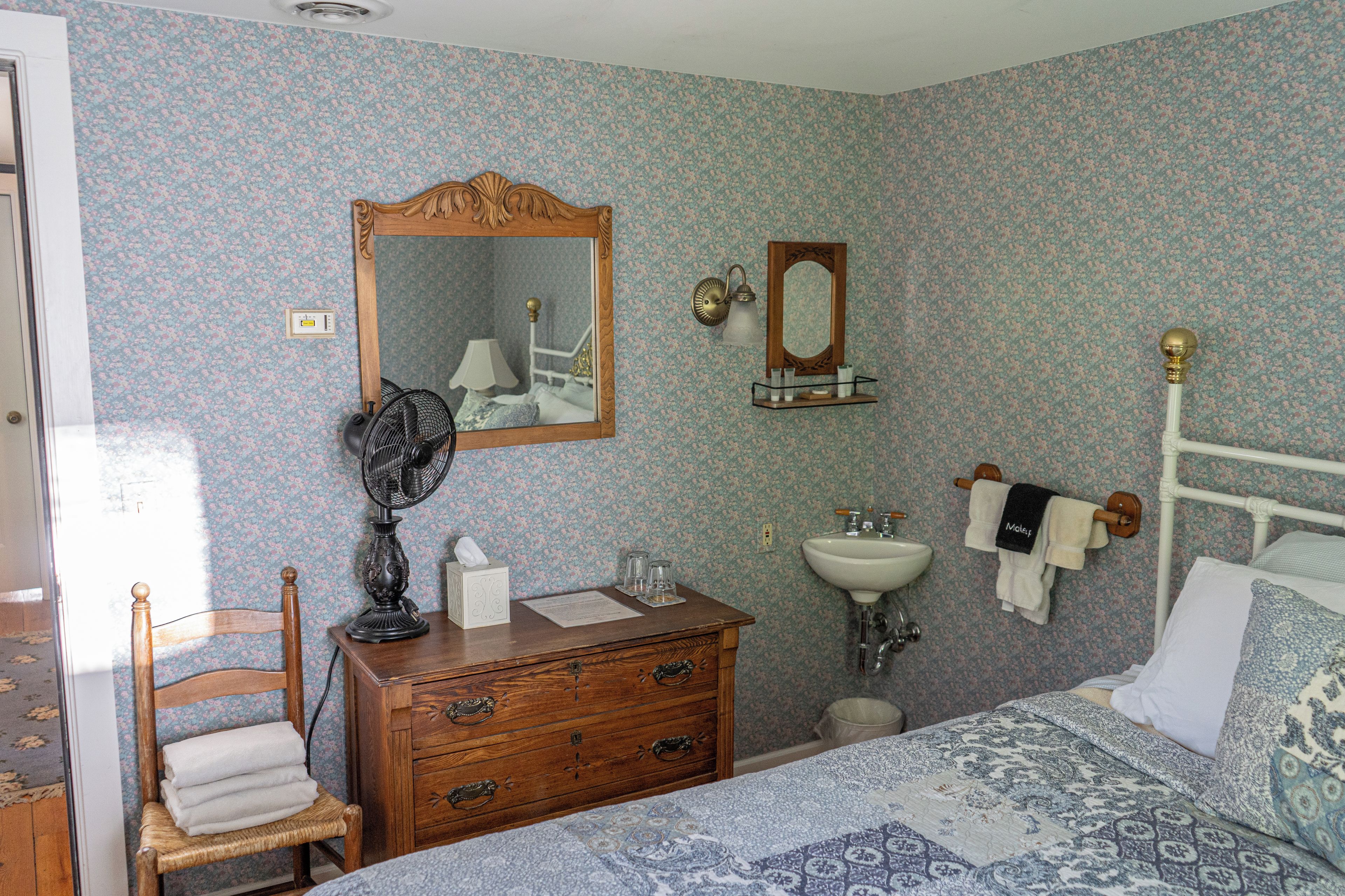 Un dormitorio con cómoda de madera, mesita de noche con lámpara, cama con ropa de cama estampada, espejo, lámpara de pared y radiador.