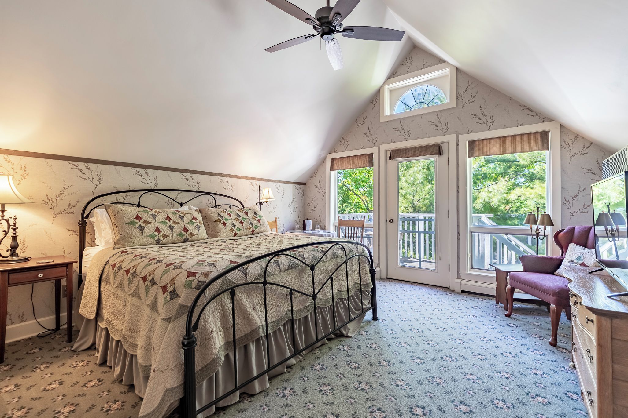 A bedroom with a bed, a desk, a chair, a ceiling fan, and a window with a view of trees.