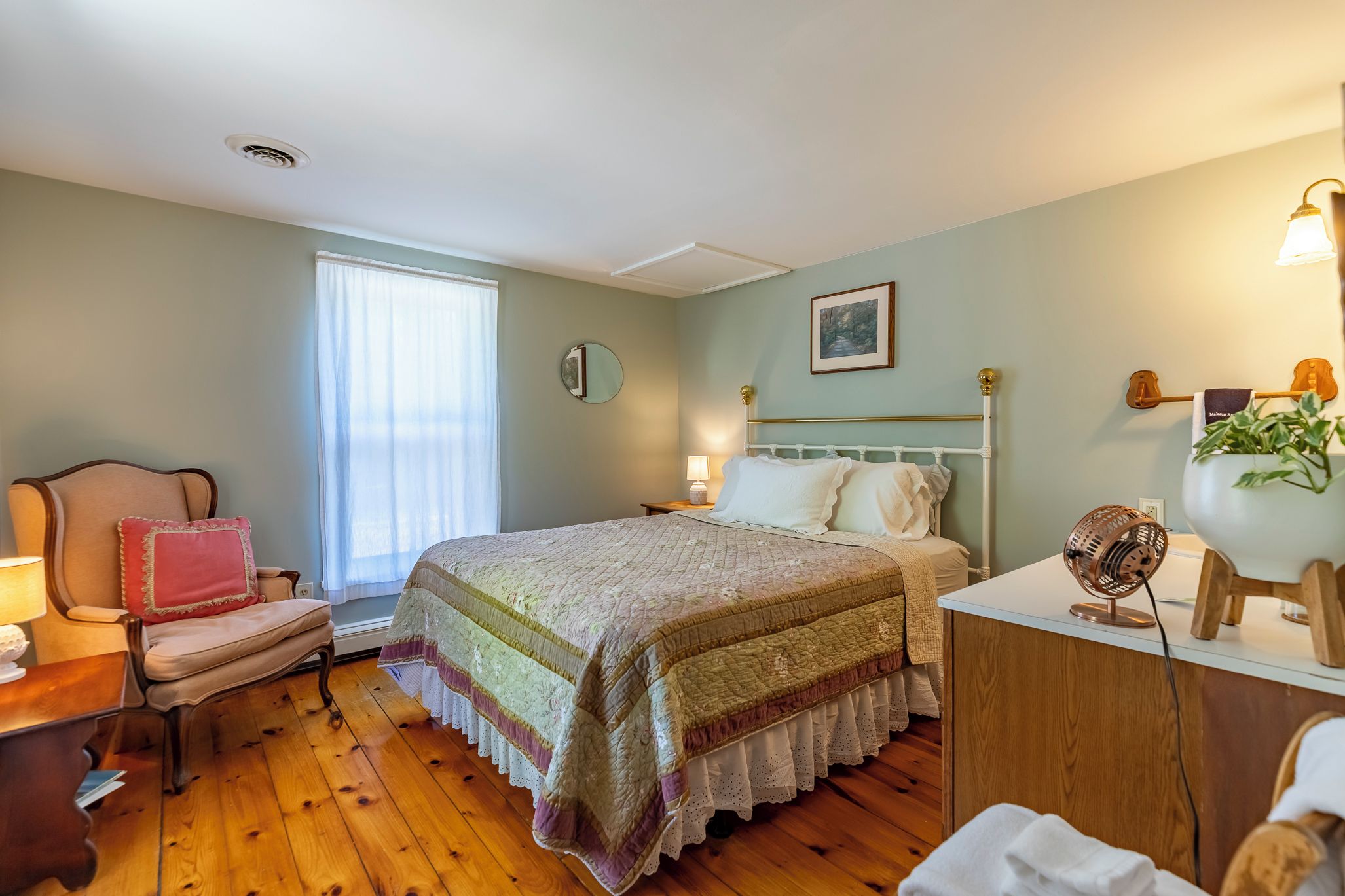 A bedroom with a bed, a chair, a bedside table, a lamp, a fan, and a window.