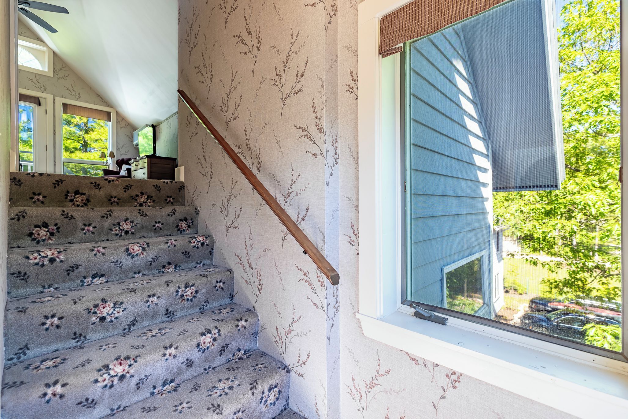 A staircase with floral carpet, a wooden handrail, and a window with a view of greenery.