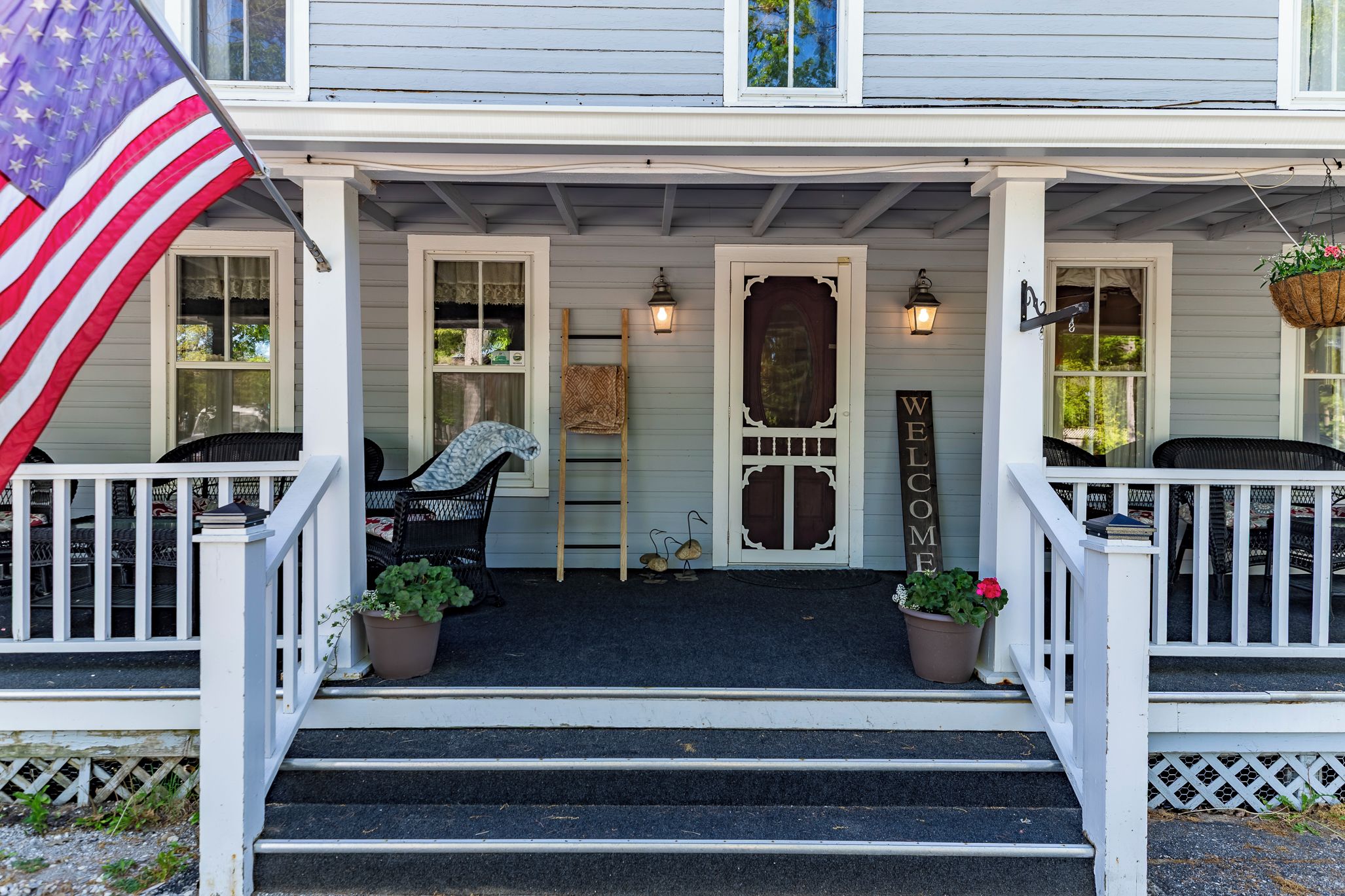 A porch with a welcome sign, steps, a flag, and potted plants.