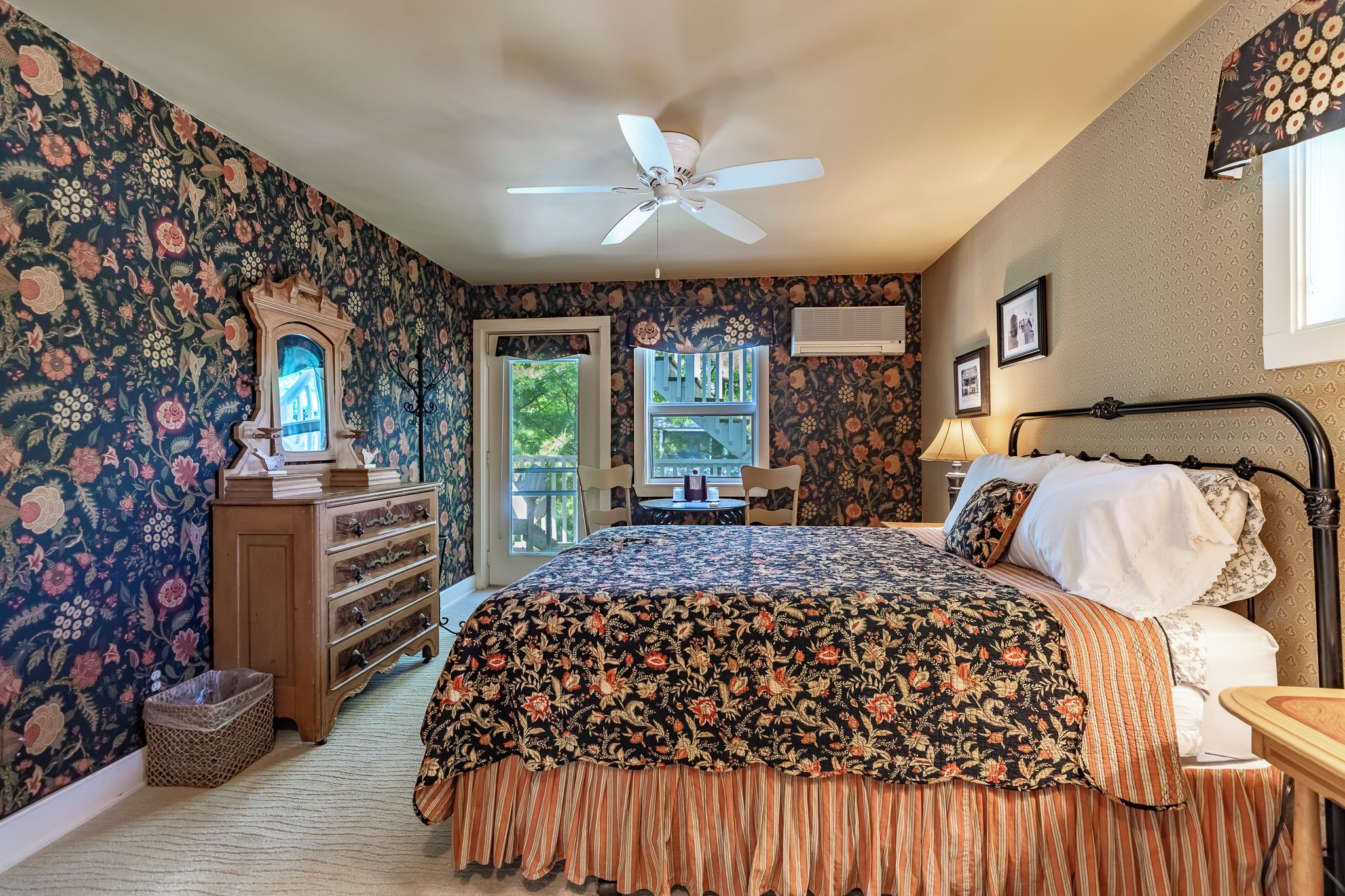 Une chambre à coucher avec une couvre-lit à motifs floraux, un ventilateur de plafond, une commode et une fenêtre donnant sur un beau paysage.