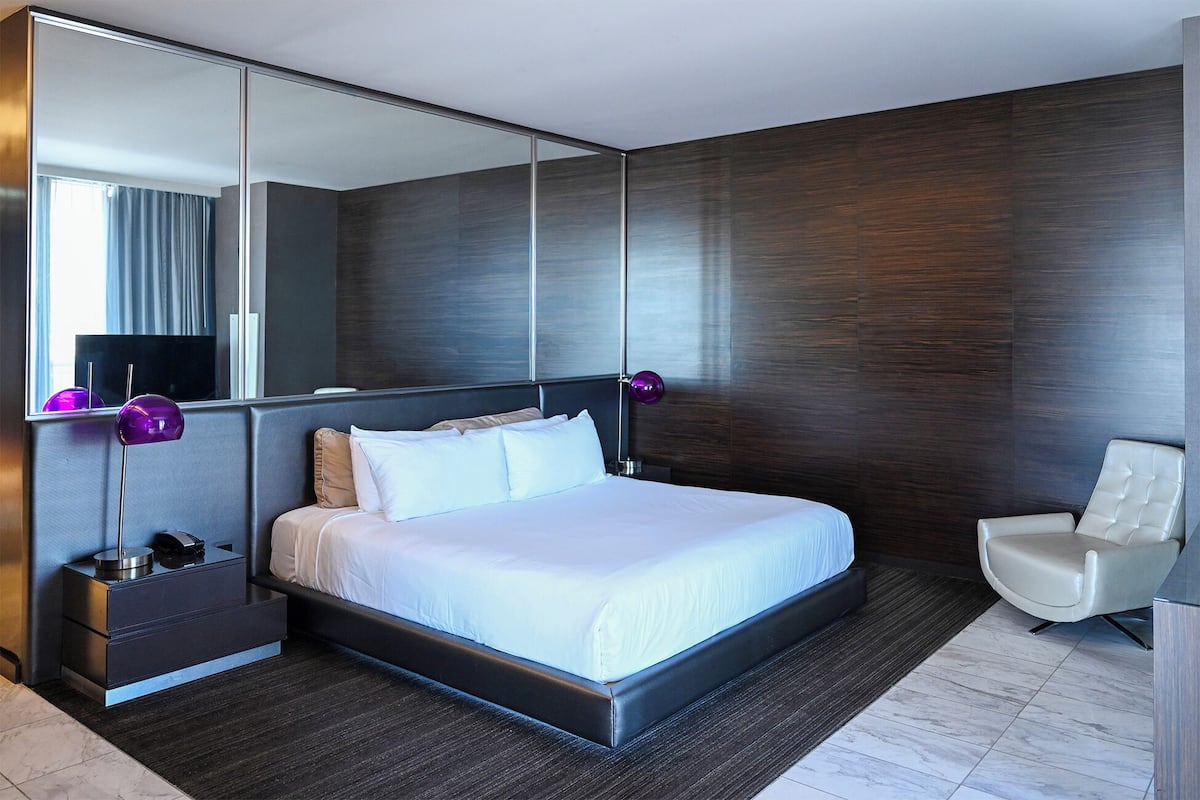 A modern hotel room with a large bed, a bedside table, a chair, and a large mirror.