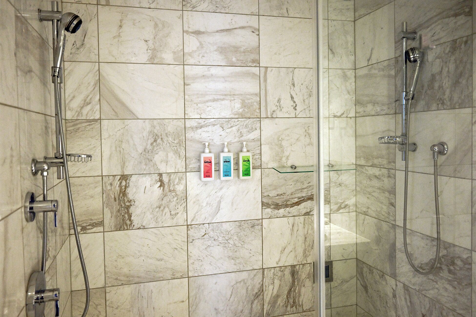 A bathroom with marble tiles, a glass shower enclosure, and three toiletries on a shelf.