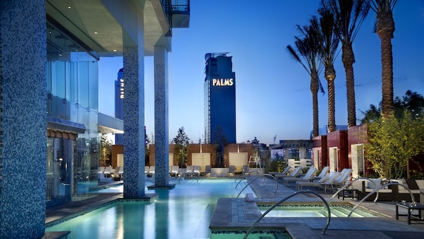 Area piscina con lettini, palme e un edificio alto con la scritta "PALMS" sullo sfondo.