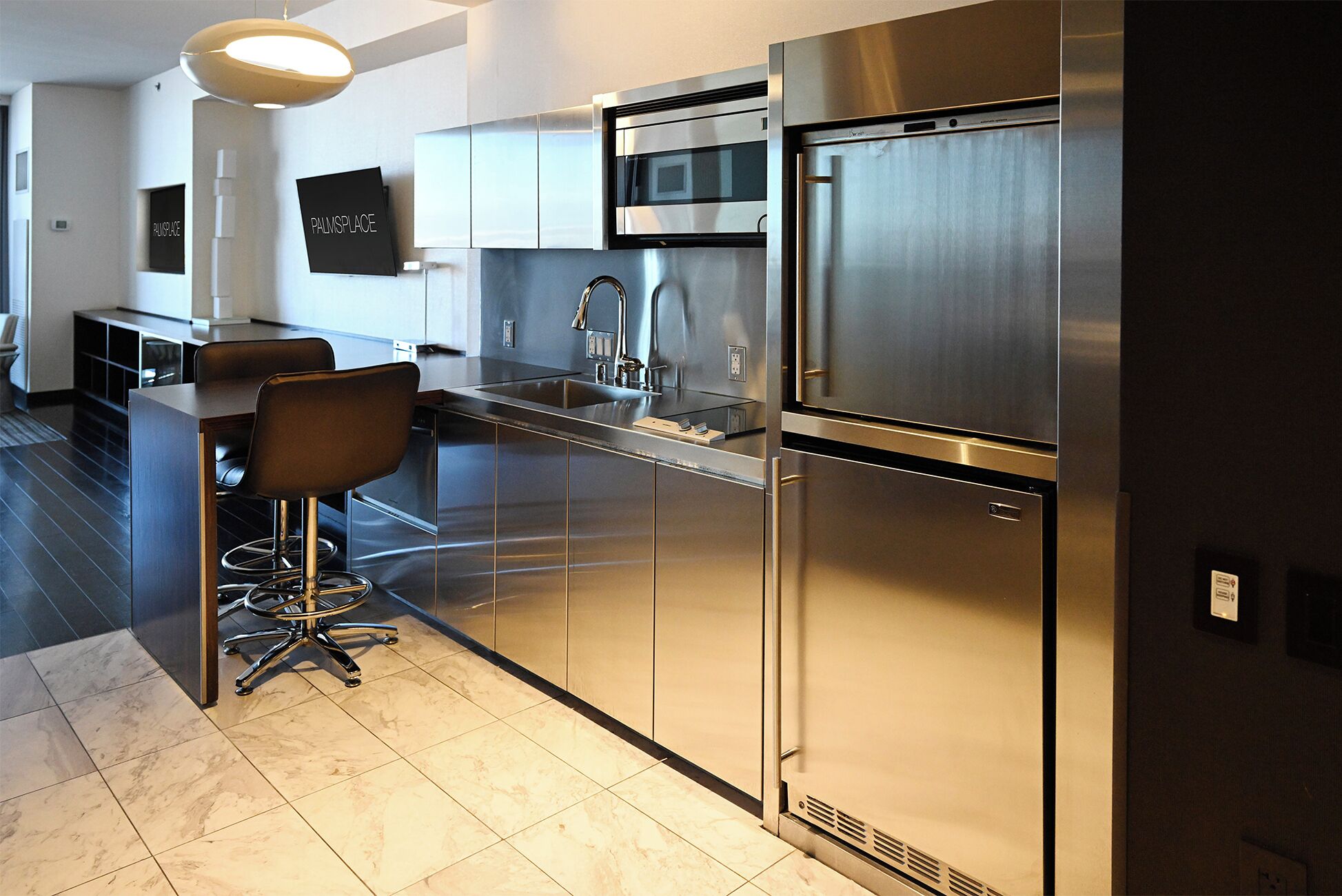 A modern kitchen with stainless steel appliances, a bar area with stools, and a visible sign reading 'PALMSPLACE'.