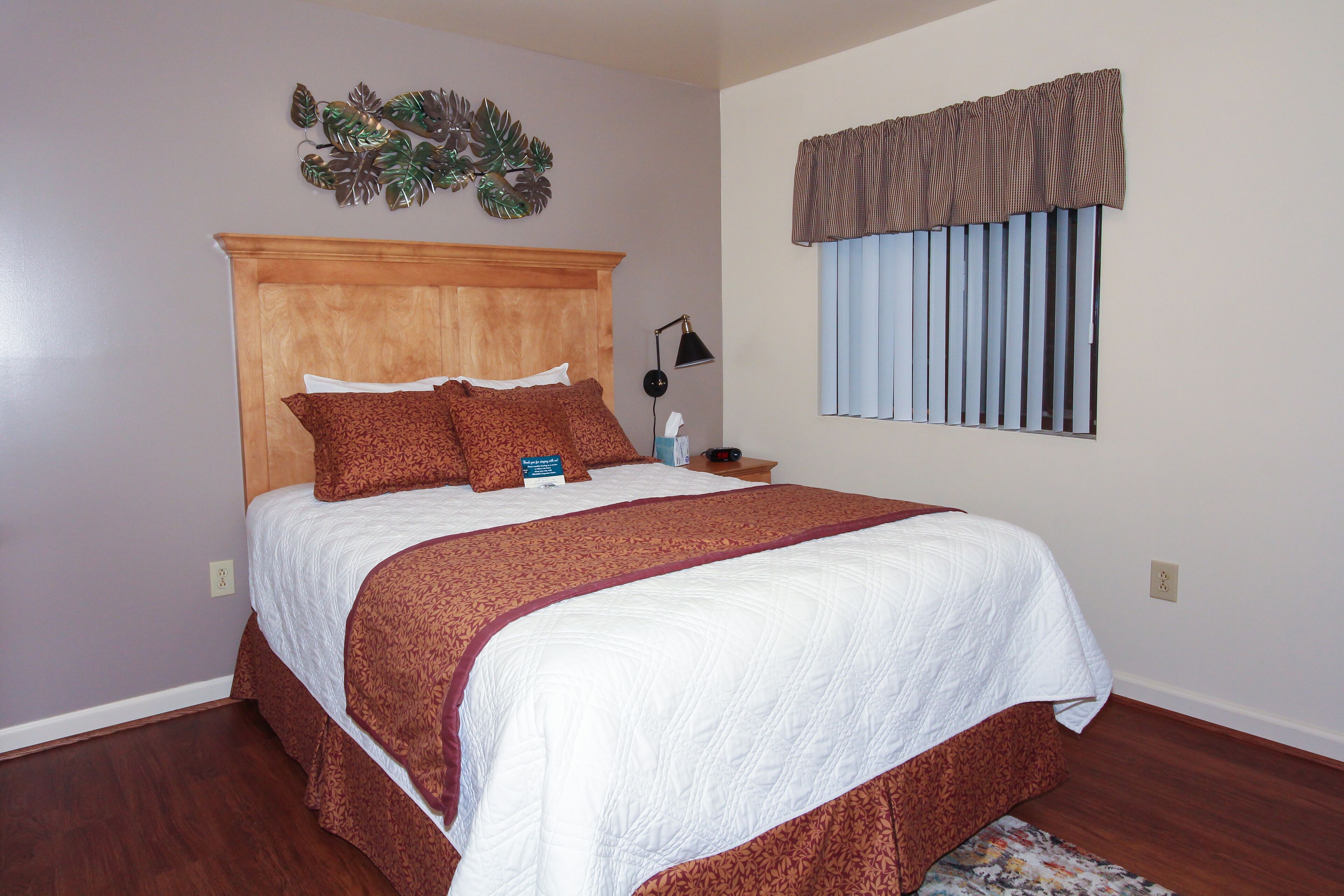 A neatly made bed with a wooden headboard, a bedside table with a lamp, and a window with blinds.
