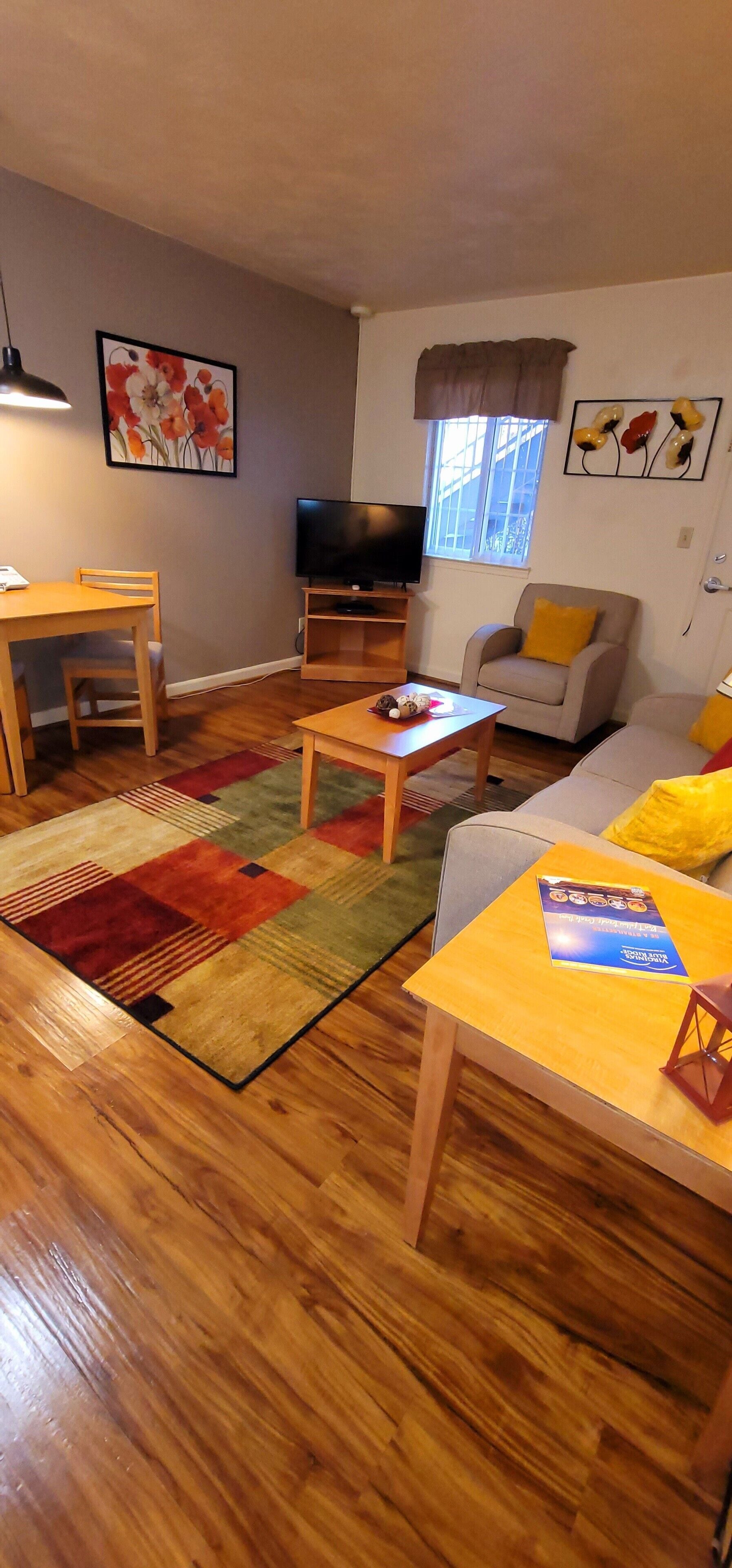 A living room with a wooden floor, a sofa, a coffee table, a TV stand, and a window with curtains.