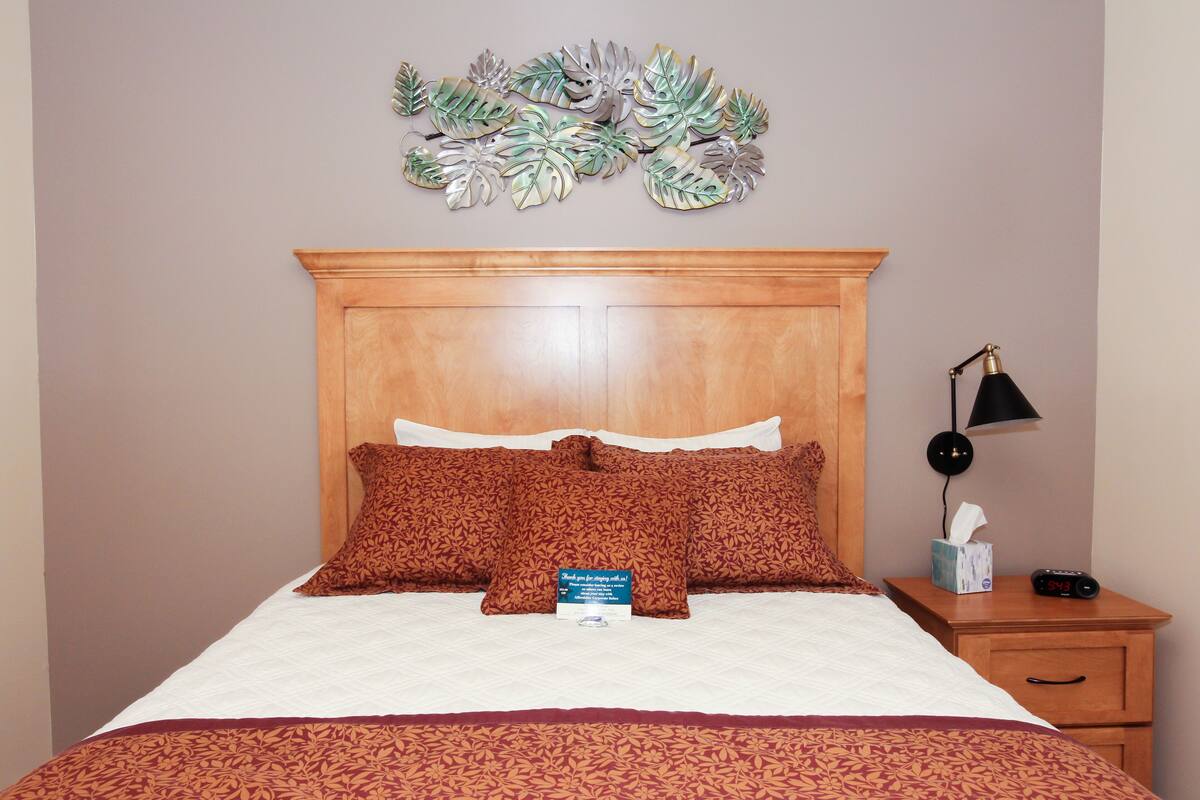A neatly made bed with patterned pillows, a wooden headboard, a bedside table with a lamp, and a wall decoration above the bed.