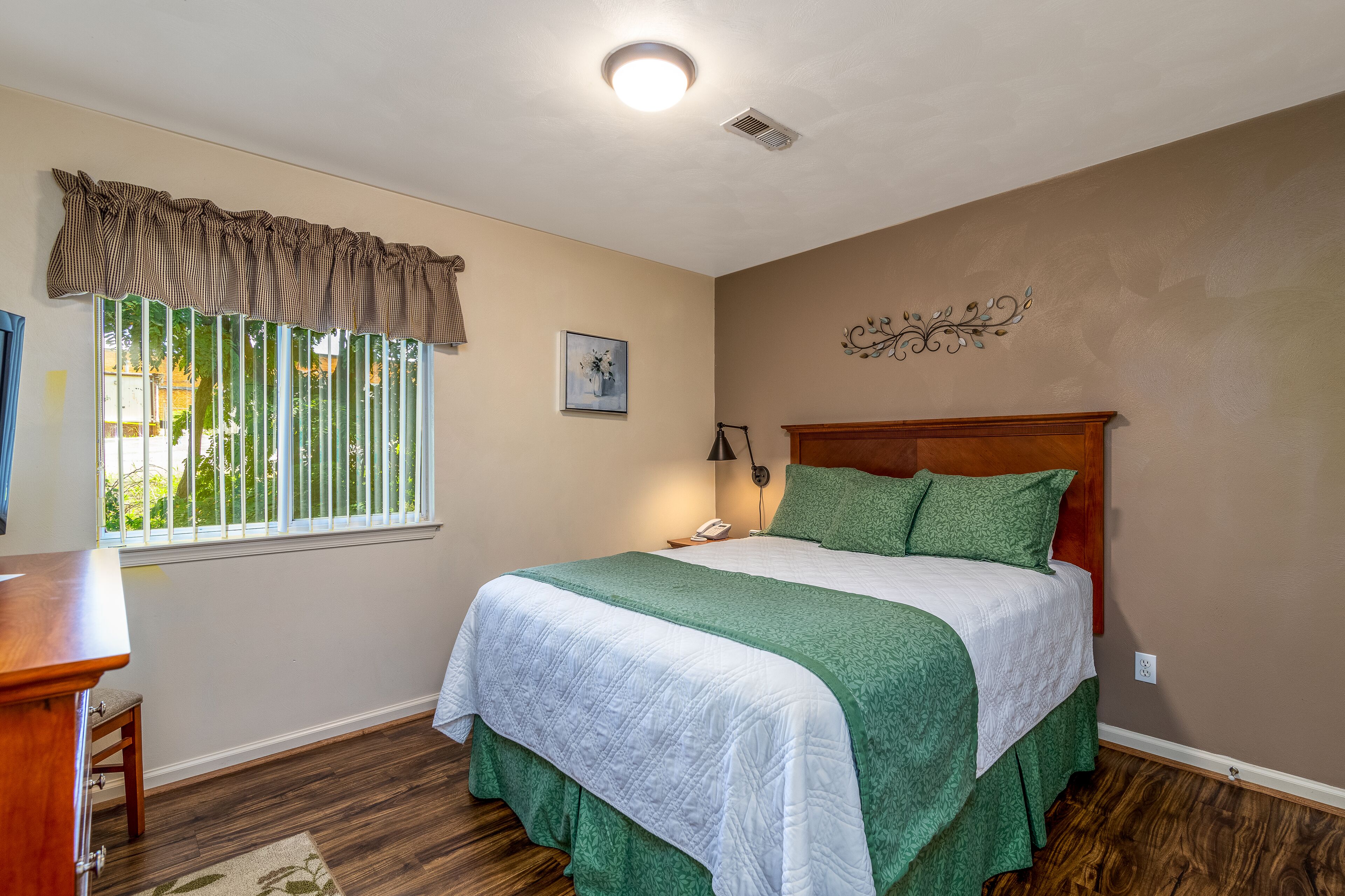 A bedroom with a large bed, a desk, a window with blinds, and a wall-mounted light fixture.