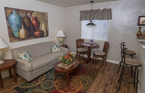 Una sala de estar con sofá, mesa de centro y sillas. Hay un cuadro en la pared, una ventana con persianas y una lámpara sobre una mesita auxiliar.