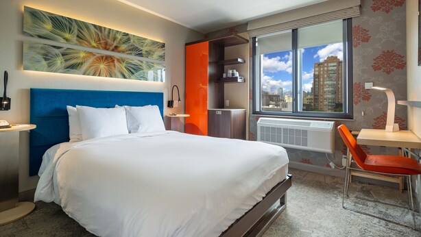 A modern hotel room with a large bed, a desk, a chair, and a window with a city view. - Hotel Le Jolie (Brooklyn)