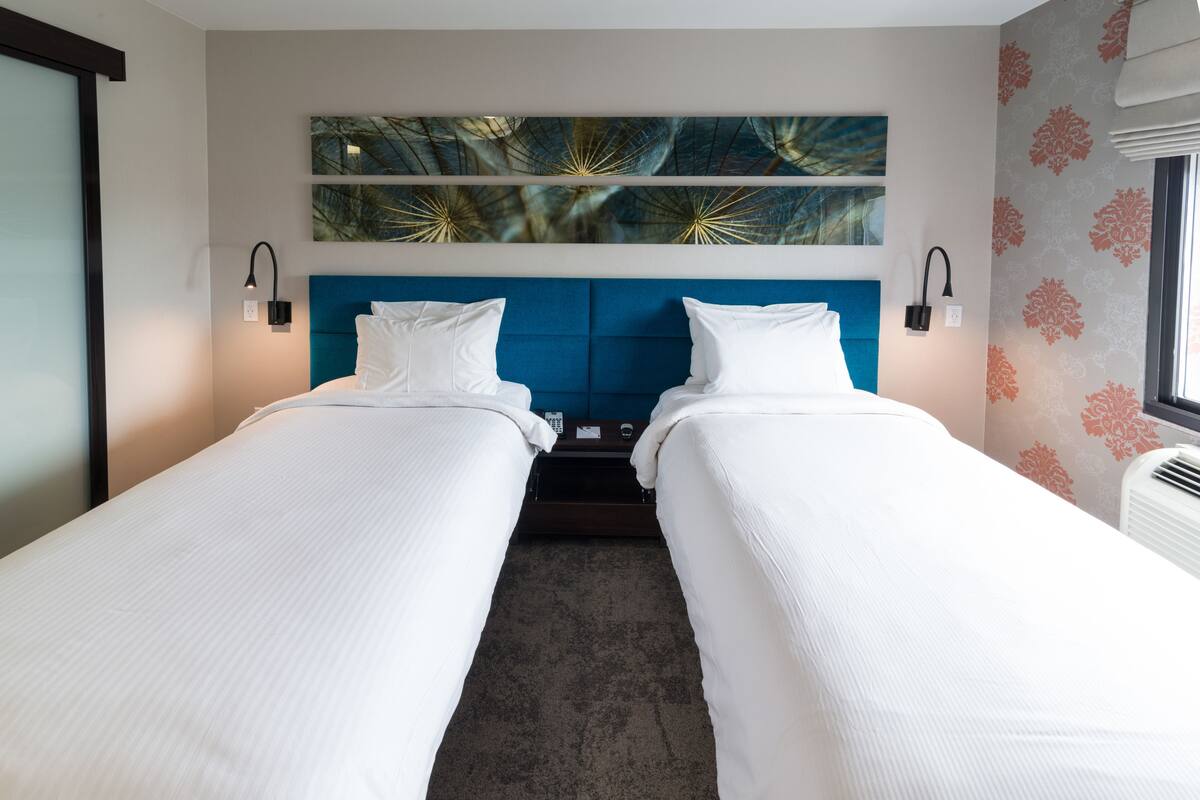 Habitación de hotel con dos camas individuales, cabecero azul, obra de arte colgada en la pared y una ventana con cortinas estampadas.