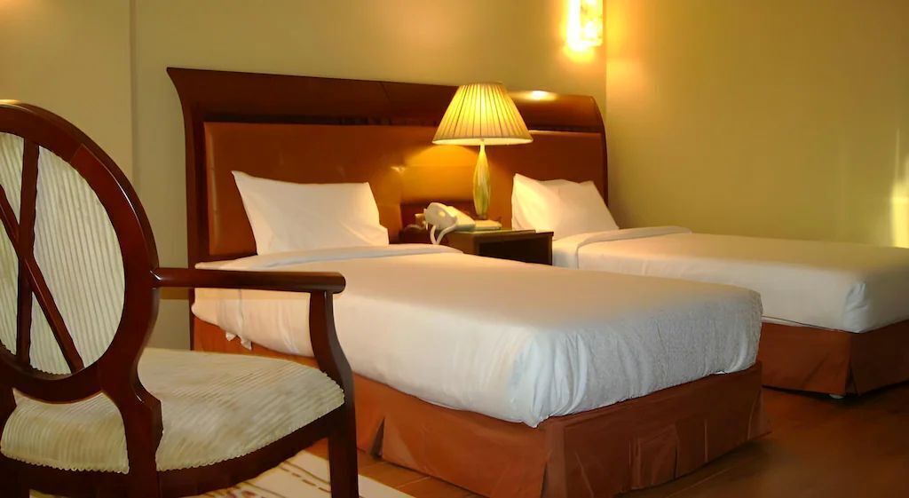 Habitación de hotel con dos camas, una silla de madera, una mesita de noche con lámpara y un teléfono.