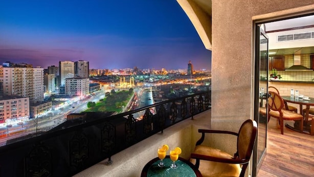 A balcony with a city view at dusk, a dining area with chairs and a table, and two drinks on a small table. - Palette Sharjah Aparthotel formerly Royal Tulip (Sharjah)