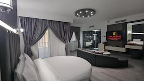 A modern hotel room with a large bed, a seating area, a TV, and a bathroom with a glass shower. - Palette Sharjah Aparthotel formerly Royal Tulip (Sharjah)