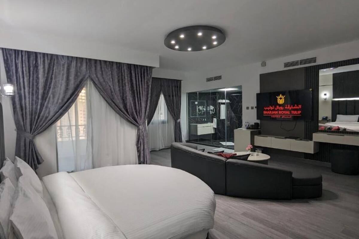 A modern hotel room with a large bed, a seating area, a TV, and a bathroom with a glass shower.