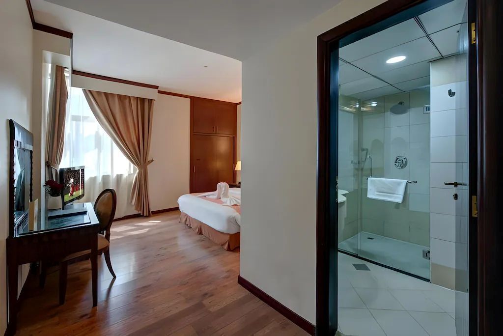 Una habitación de hotel con piso de madera, una cama, un escritorio con una silla, cabina de ducha de vidrio y una ventana con cortinas.