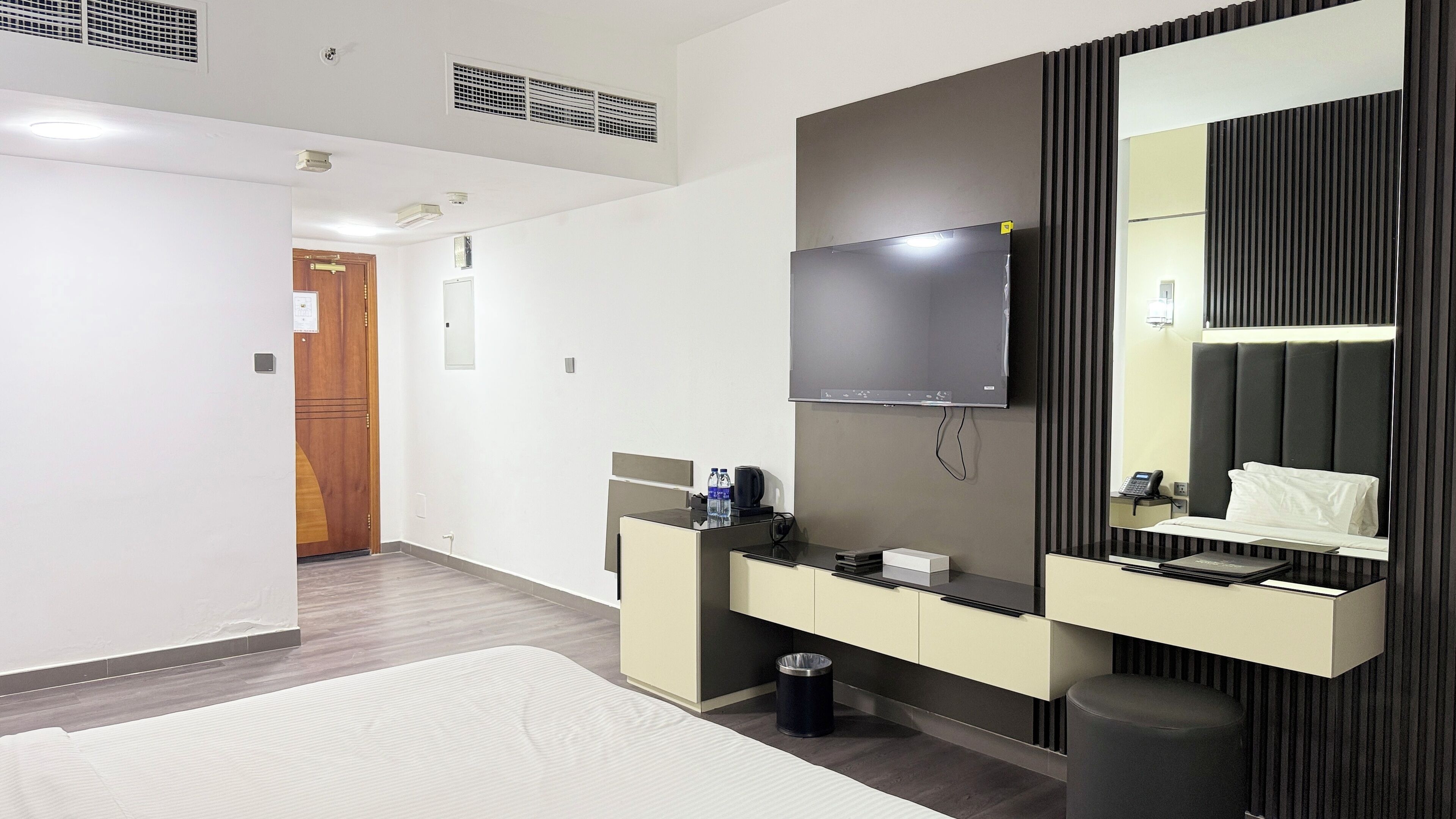Une chambre d’hôtel moderne avec un grand lit, un téléviseur fixé au mur, un petit bureau avec une bouteille d’eau et une poubelle.