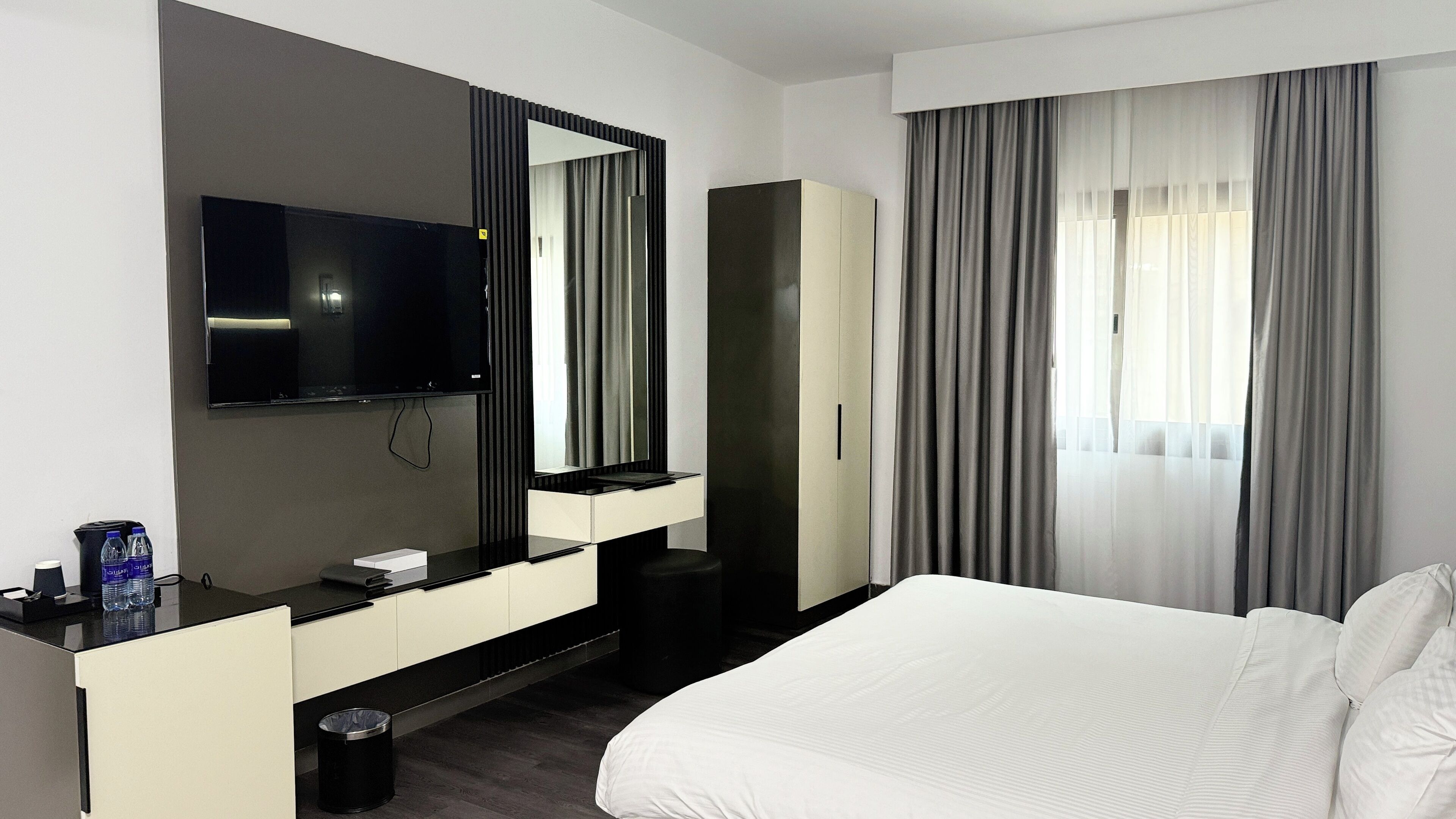 Ett modernt hotellrum med en stor säng, en platt-TV som är monterad på väggen, en minibar och ett stort fönster med gardiner.