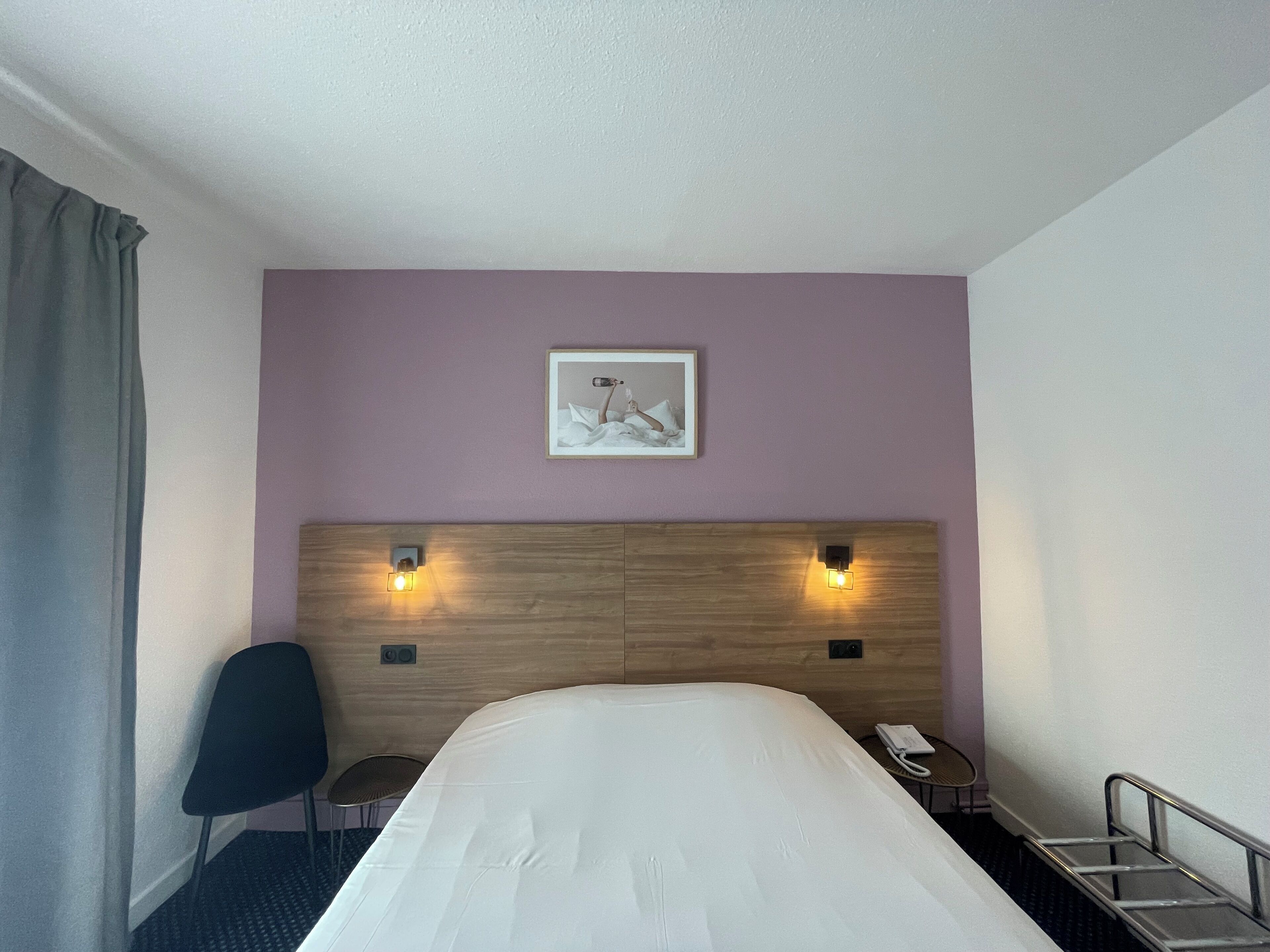 Habitación de hotel con una cama, una silla, una mesita, un teléfono y un cuadro en la pared.